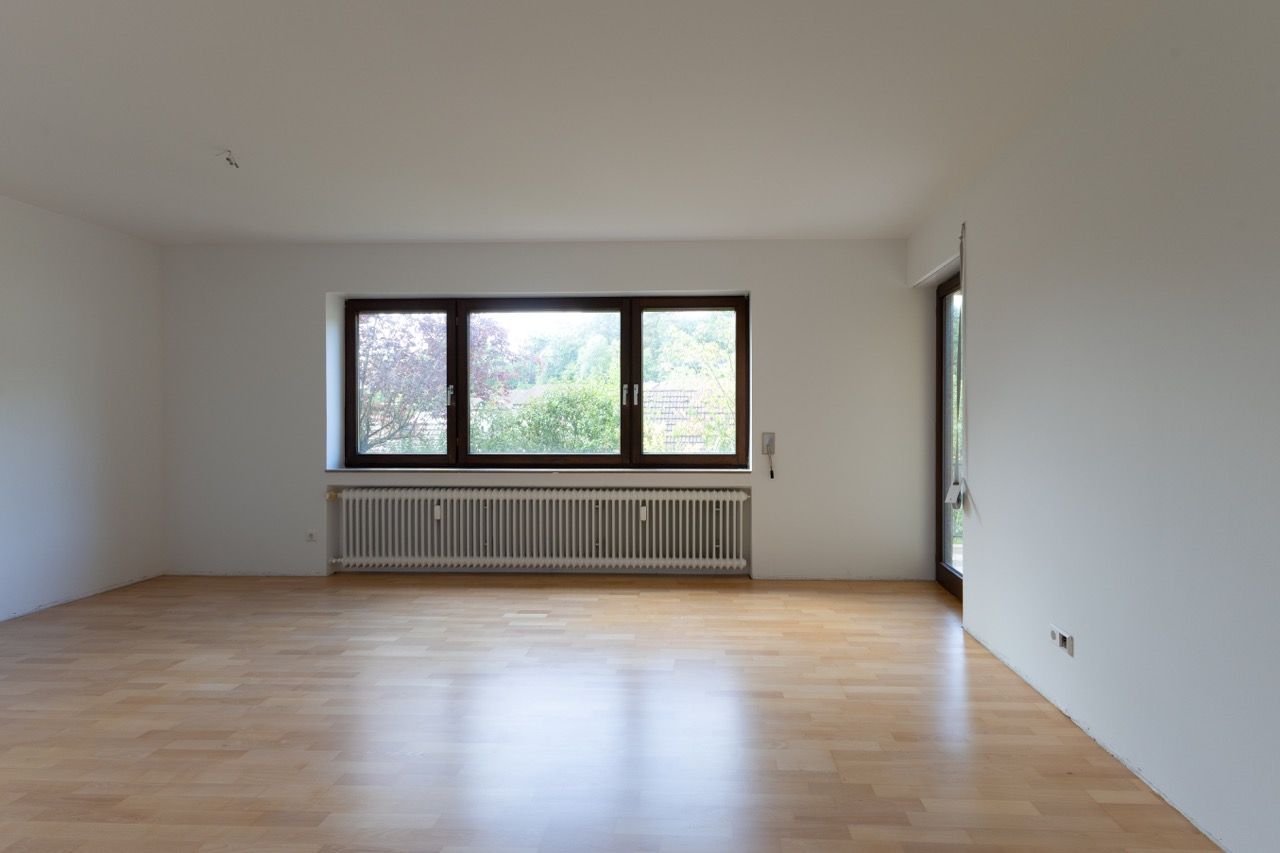 Leeres Wohnzimmer mit Holzboden, weißen Wänden und einem großen Fenster über einem Heizkörper.