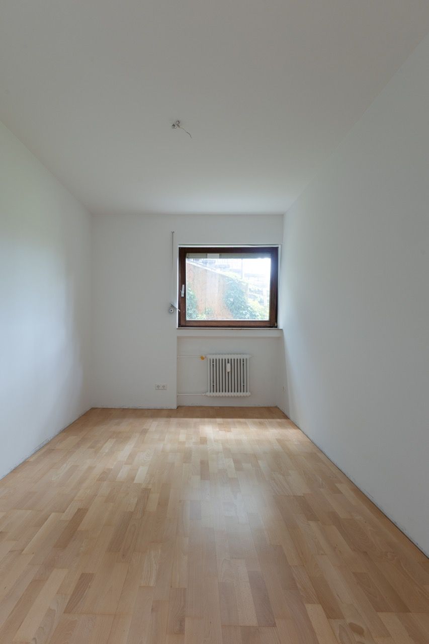 Leerer weißer Raum mit hellem Holzboden, kleinem Fenster und Heizkörper an der gegenüberliegenden Wand.