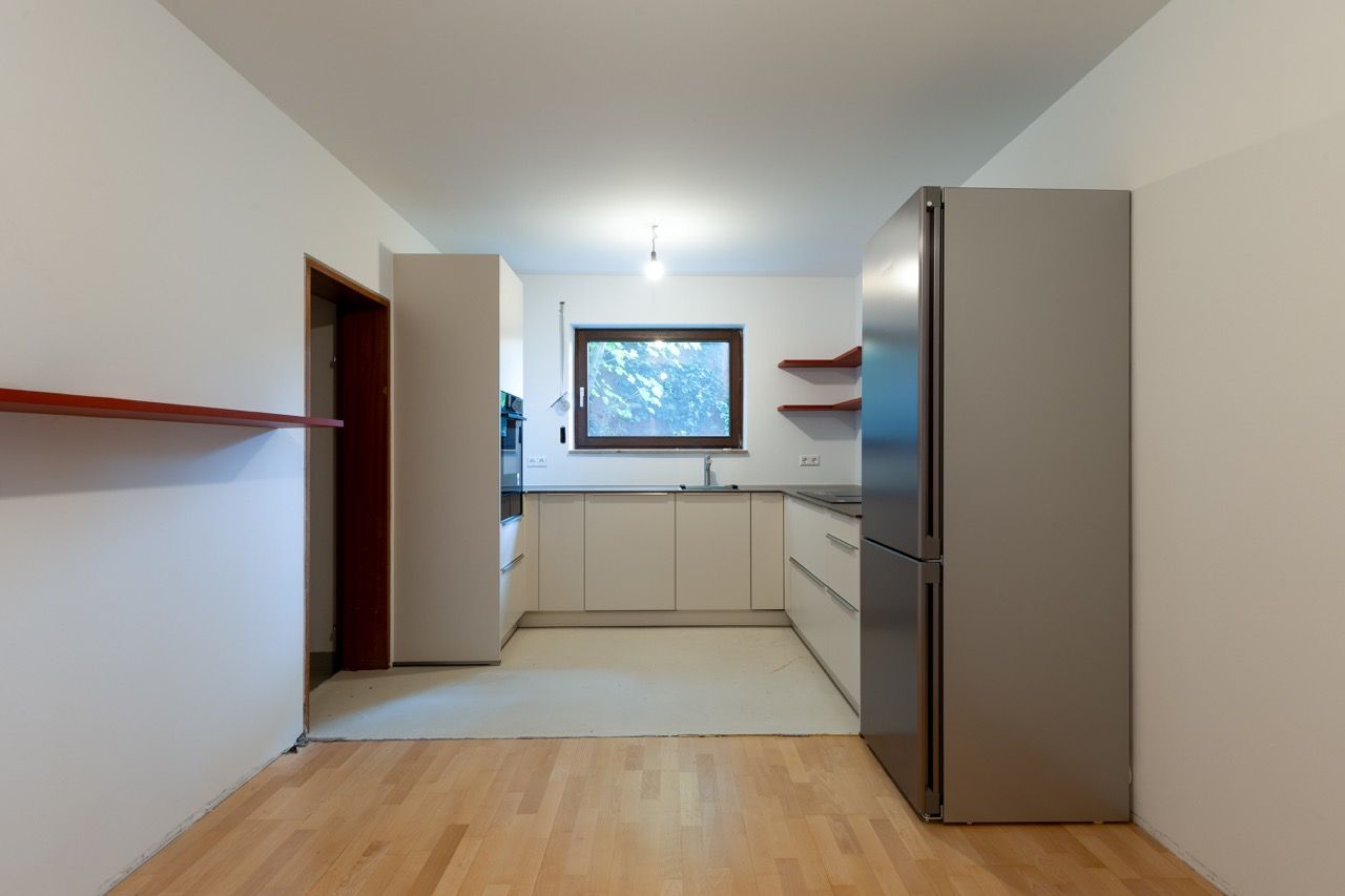 Leere, moderne Küche mit hellem Holzboden, weißen Schränken, Edelstahlkühlschrank und einem Fenster über der Spüle