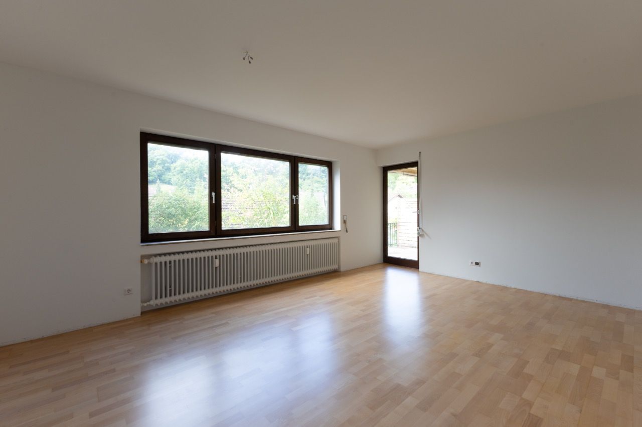 Leeres Wohnzimmer mit Holzboden, großen Fenstern, Heizkörper und offener Tür zum Balkon