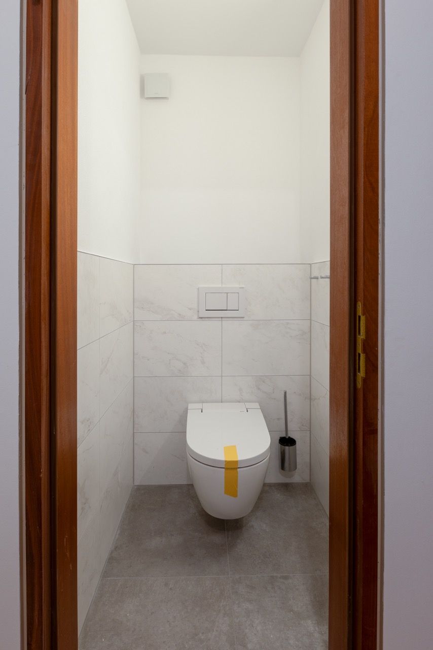 Kleines weißes Badezimmer mit wandhängender Toilette und grauem Fliesenboden, gesehen durch eine offene Tür.