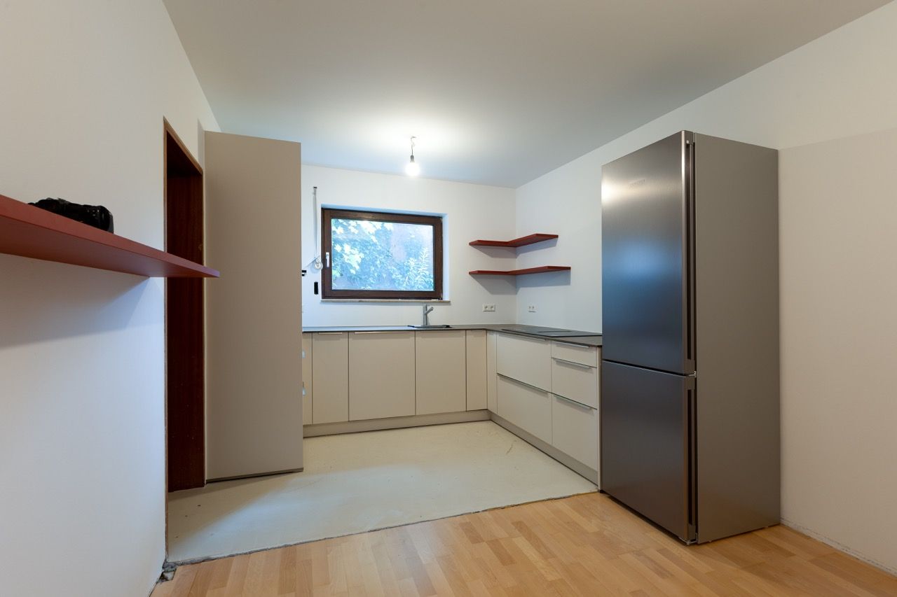 Moderne, minimalistische Küche mit hellen Schränken, Holzboden, Edelstahlkühlschrank und einem Fenster über der Arbeitsplatte.