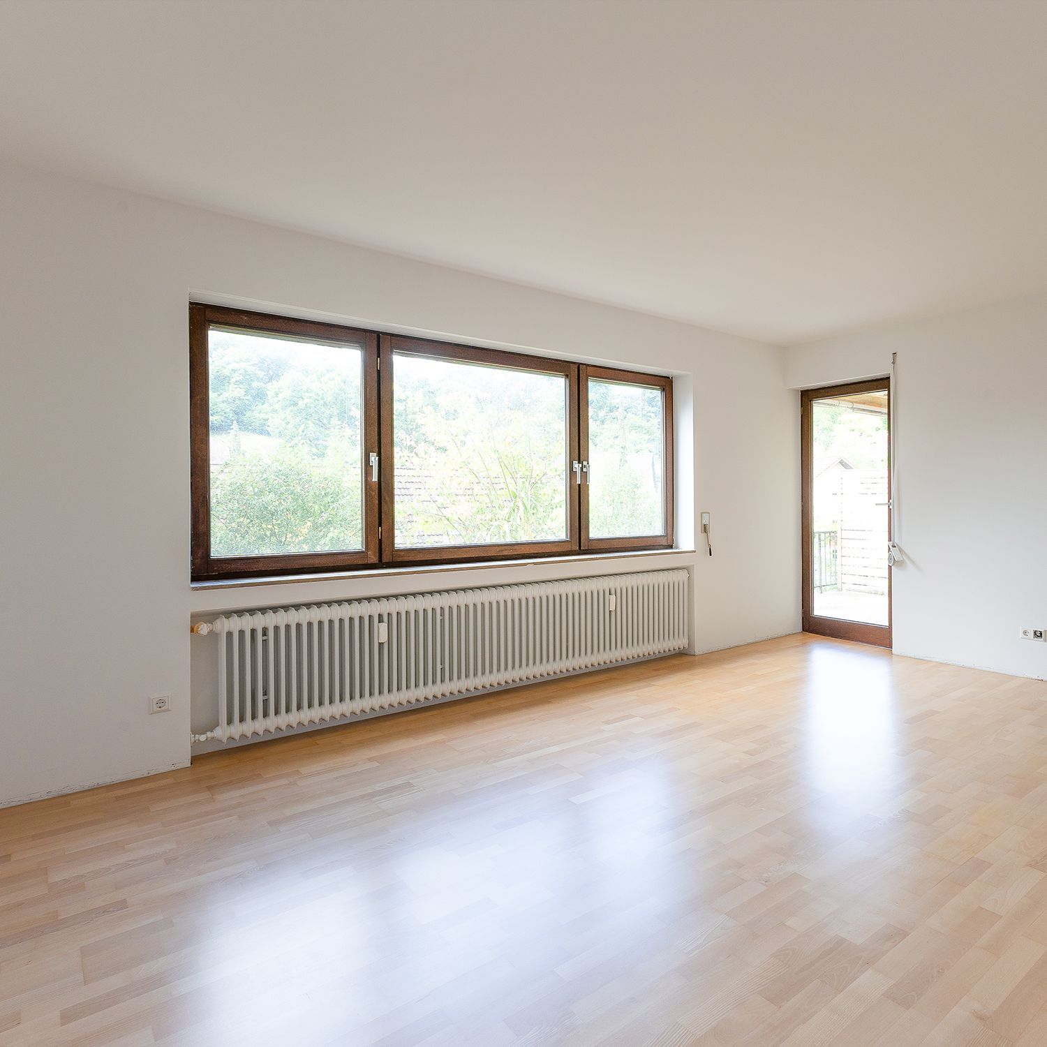 Heller, leerer Raum mit Holzboden, großen Fenstern, Heizkörper und Glastür.