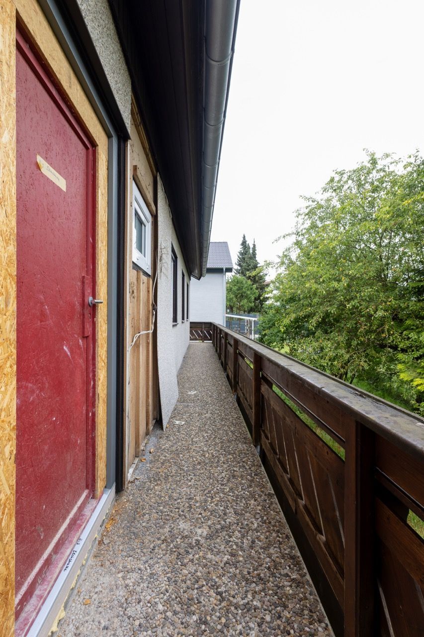 Schmaler Kiesbalkon neben einer roten Tür und einem Holzgeländer mit Blick auf grüne Bäume und Wasser.