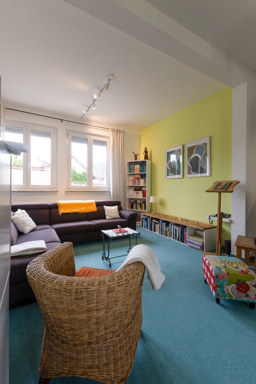 Helles Wohnzimmer mit türkisfarbenem Teppich, Korbsessel, dunklem Sofa, gelber Wand und großen Fenstern.