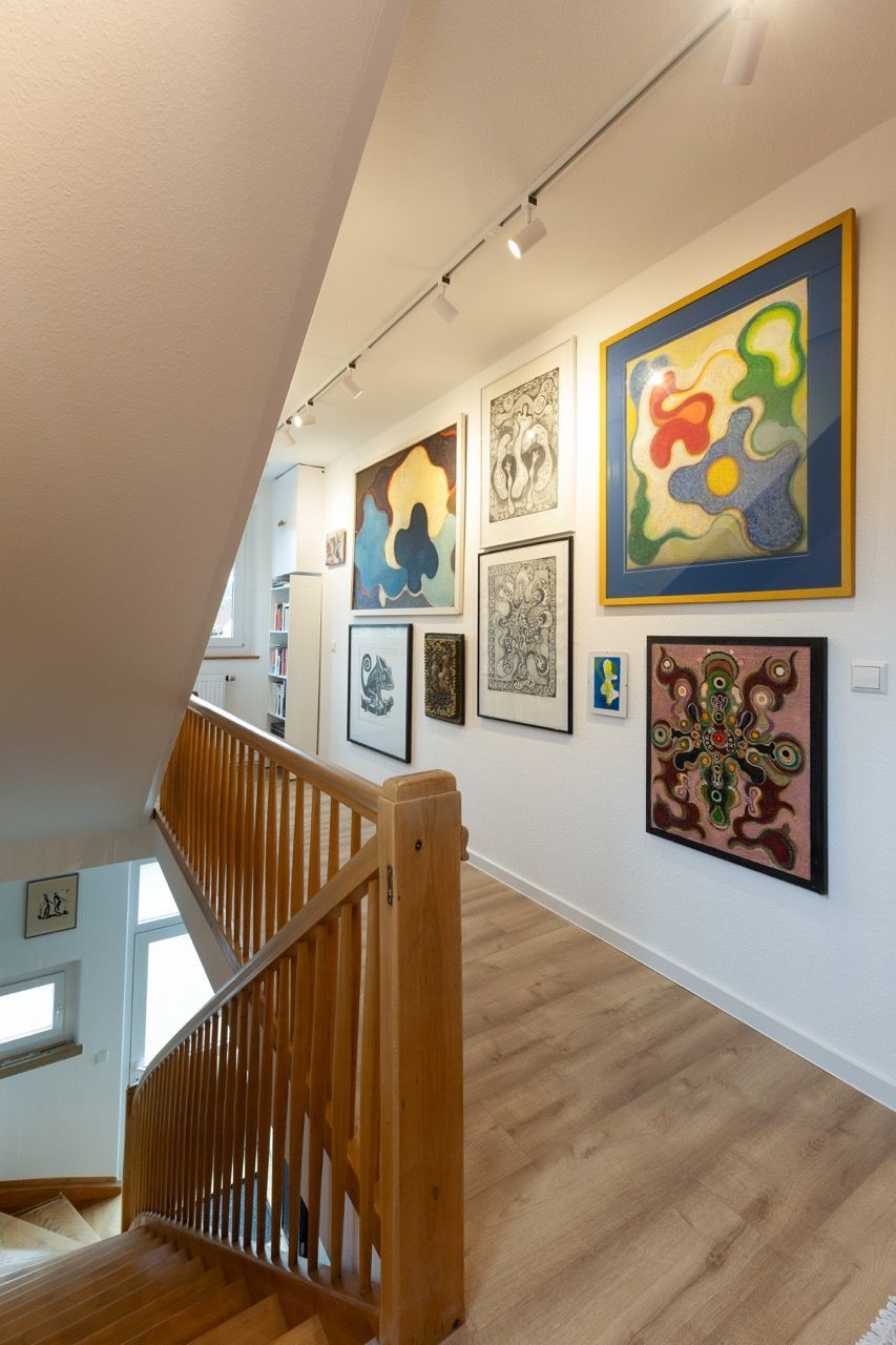 Flurgalerie mit farbenfrohen, gerahmten Gemälden an einer weißen Wand und einem Holzgeländer neben einer Treppe