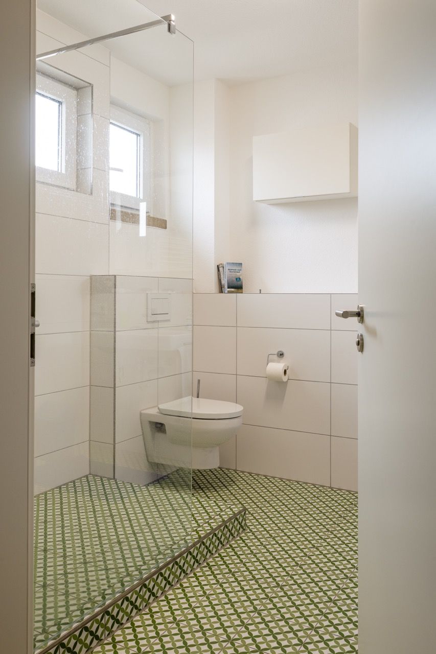 Kleines weißes Badezimmer mit grün gemustertem Fliesenboden, wandhängendem WC, Dusche und Fenster.