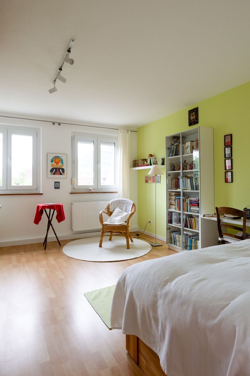 Helles Schlafzimmer mit lindgrüner Wand, Bett, Stuhl, Bücherregal und zwei Fenstern auf Holzboden.