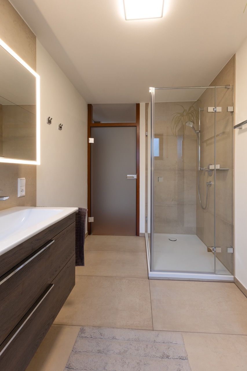 Modernes Badezimmer mit Doppelwaschtisch, großem Spiegel und verglaster Dusche auf beigefarbenem Fliesenboden.