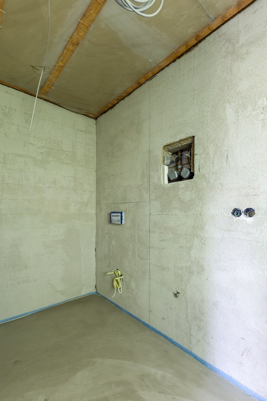 Leerer, unfertiger Raum mit Betonwänden, freiliegender Decke und Sanitär-/Elektroinstallationen.