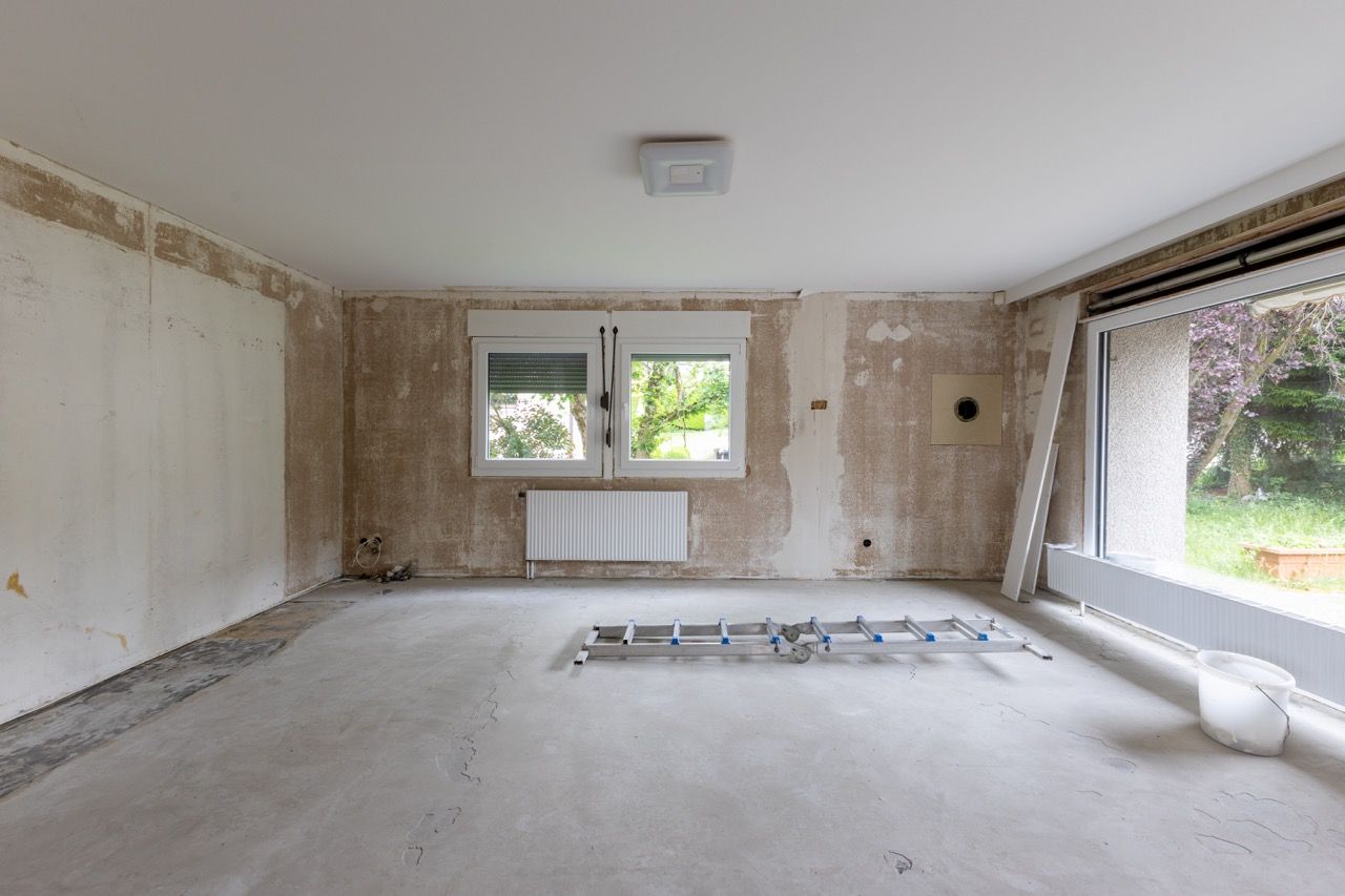 Leeres Zimmer im Renovierungszustand mit kahlen Wänden, Betonboden, Fenster, Heizkörper und freiliegenden Kabeln