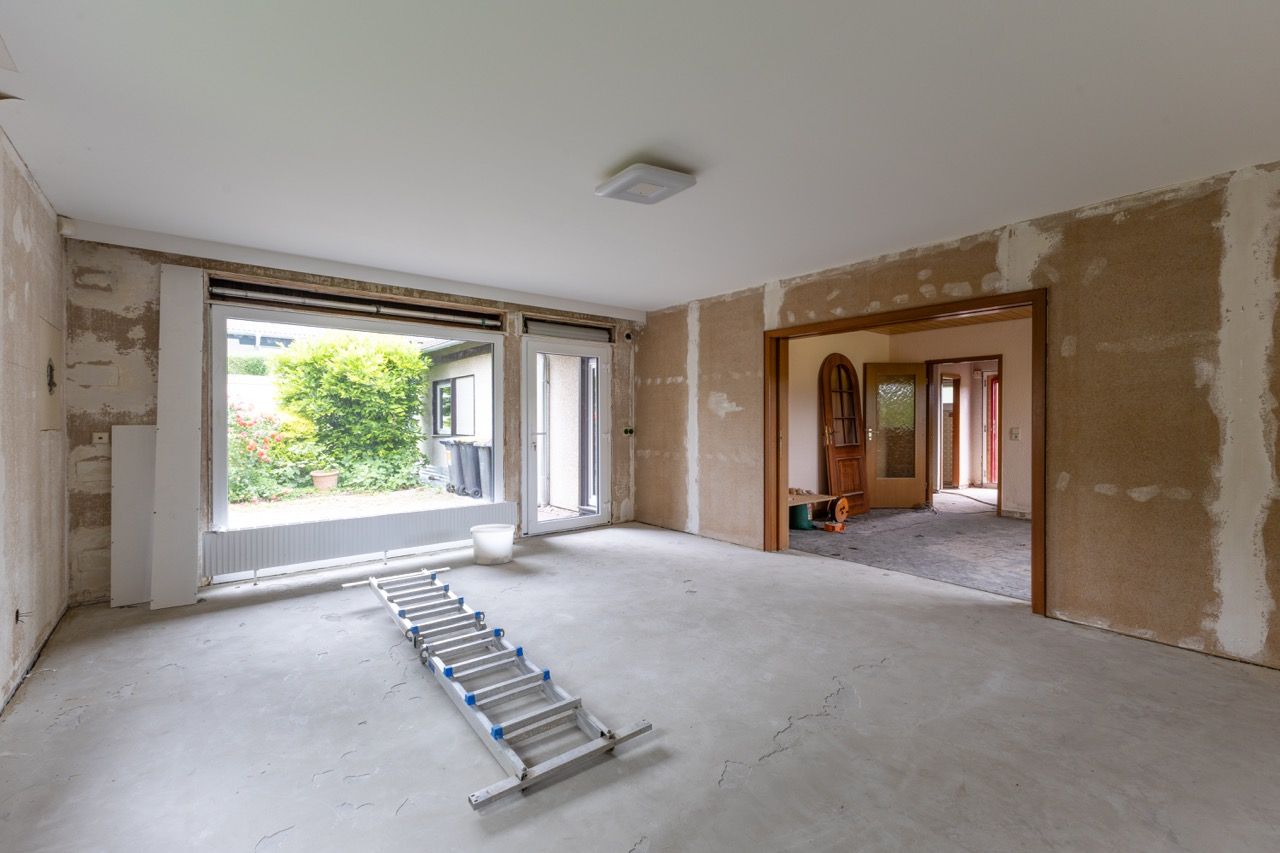 Leeres, unfertiges Wohnzimmer mit freiliegenden Gipskartonwänden, großem Fenster und einer Leiter auf dem Boden