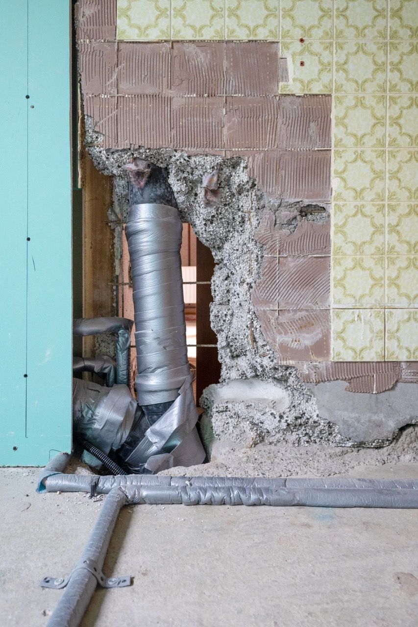 Beschädigte Wand mit freiliegendem silbernen Lüftungsrohr neben türkisfarbener und gelber Tapete in einem Raum, der renoviert wird.