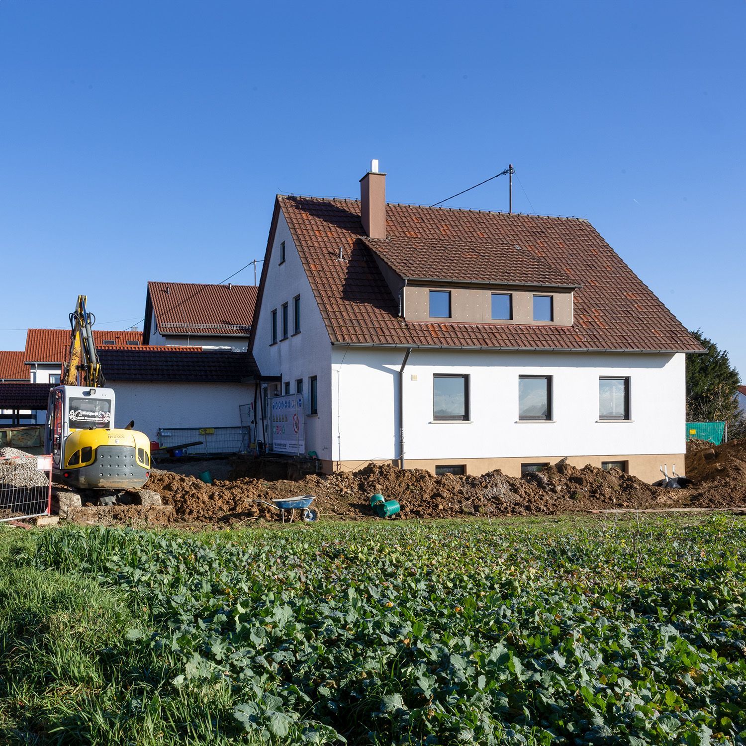 Ein im Bau befindliches Haus neben einem Feld, mit Bagger und Arbeitern unter klarem blauen Himmel