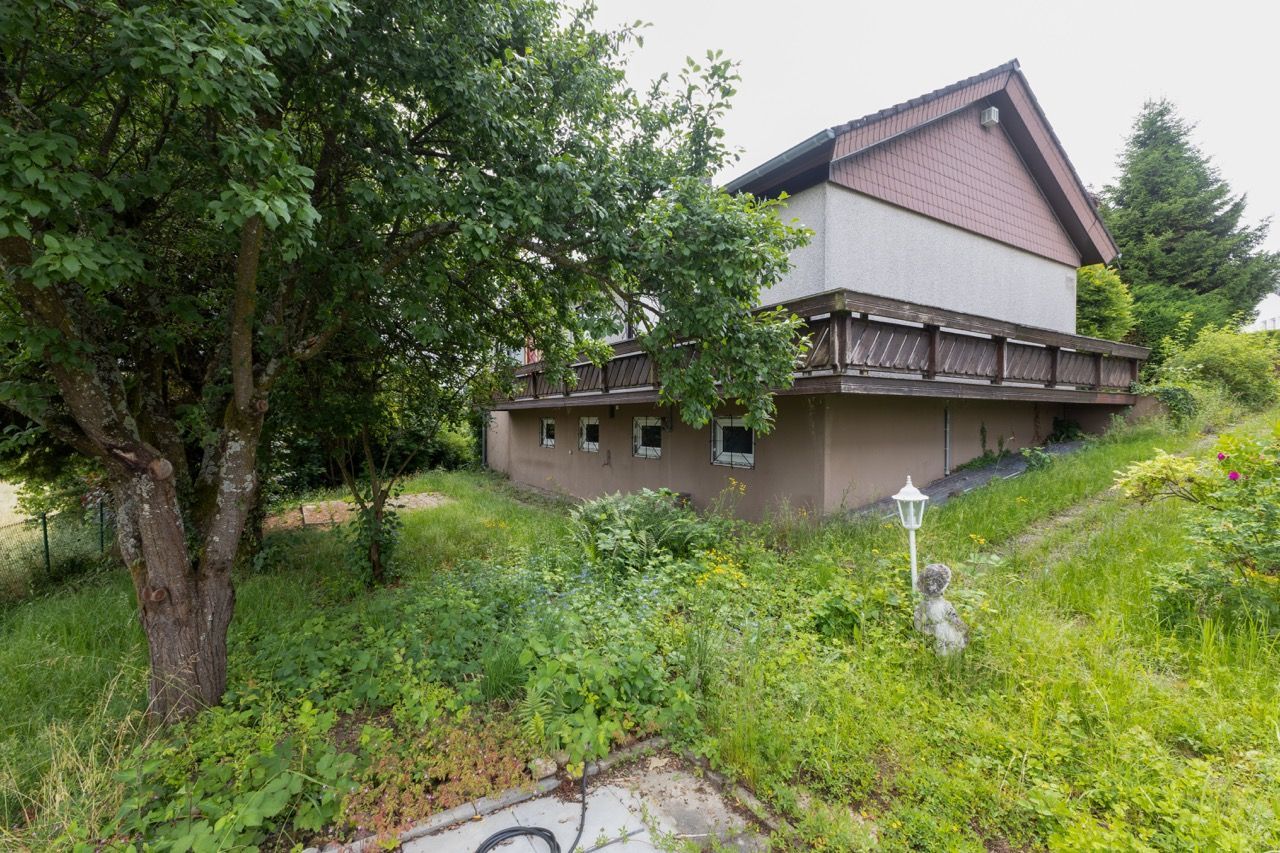 Kleines Haus mit Balkon an einem grasbewachsenen Hang, umgeben von Bäumen und üppigem Grün.