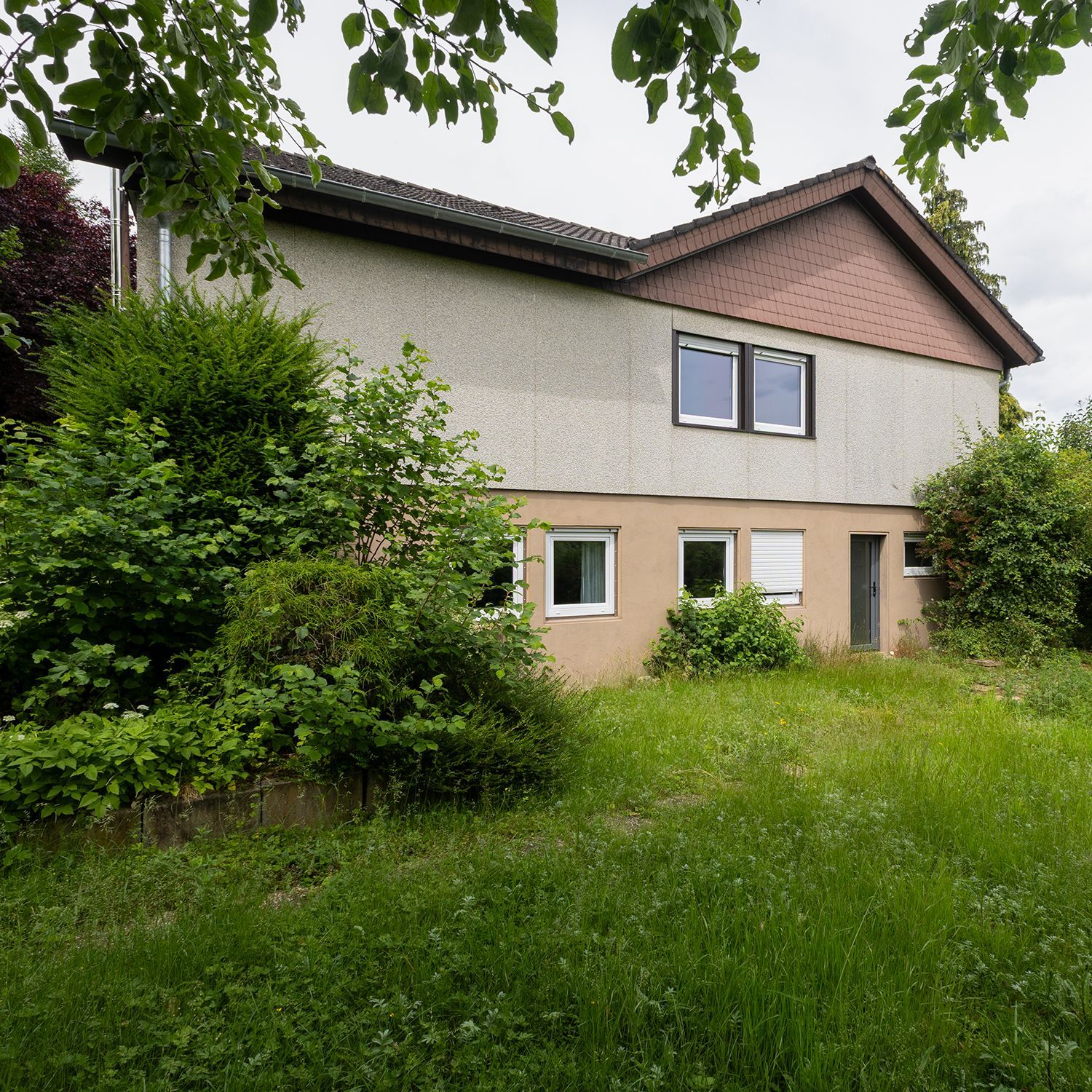 Verwilderter Vorgarten vor einem beigefarbenen zweistöckigen Haus mit Bäumen und Sträuchern.