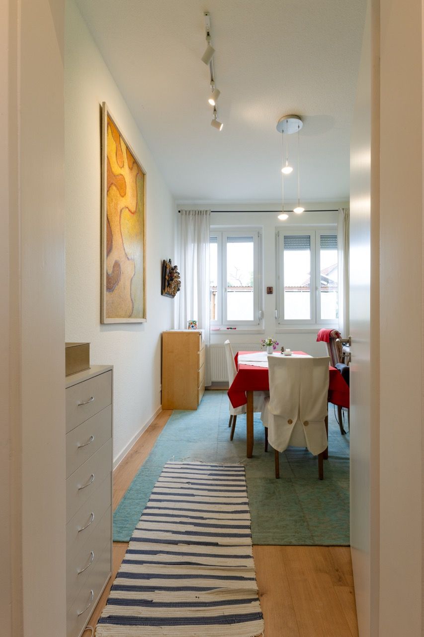 Ein heller Flur führt zu einem kleinen Essbereich mit roter Tischdecke, gestreiftem Tischläufer und großem Fenster.