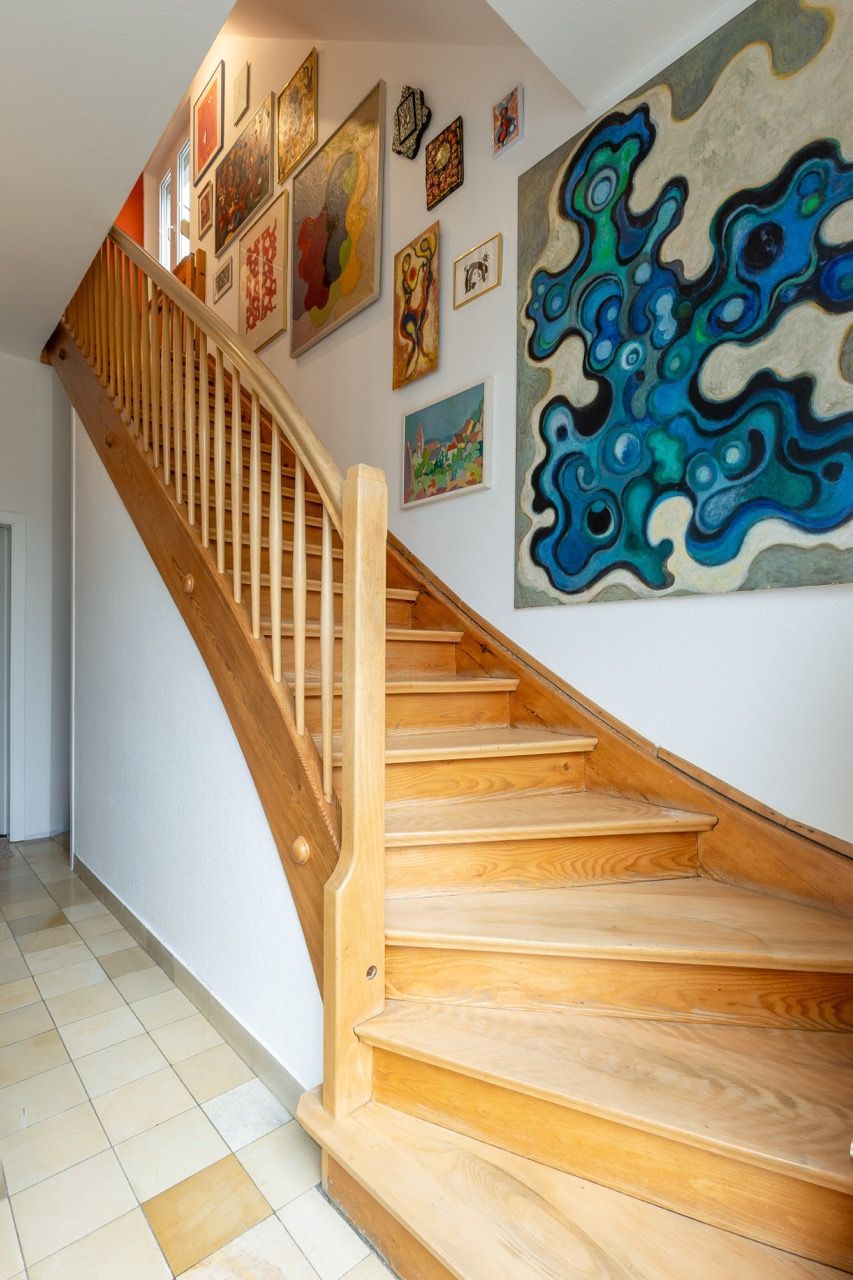 Eine Holztreppe in einem Flur, an dessen Wänden gerahmte Kunstwerke und ein blaues abstraktes Gemälde hängen.