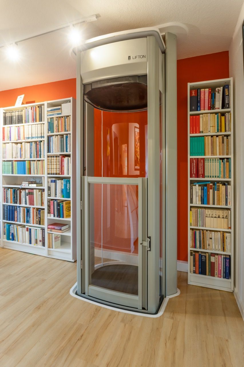 Ein moderner Aufzug in einem gemütlichen Bibliotheksraum mit weißen Bücherregalen und warmen orangefarbenen Wänden
