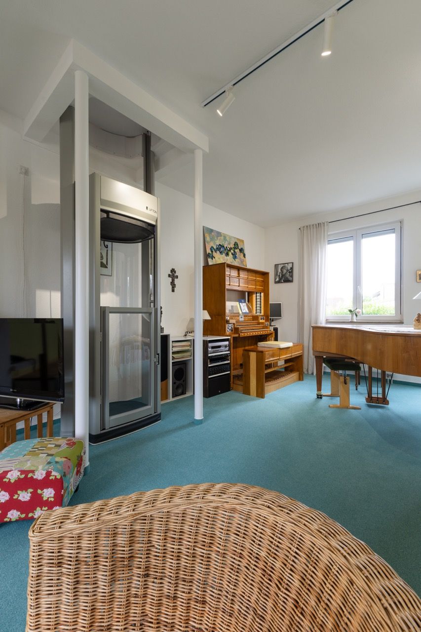 Helles Zimmer mit Korbstühlen, türkisfarbenem Teppich, einem Klavier und einem großen Fenster.
