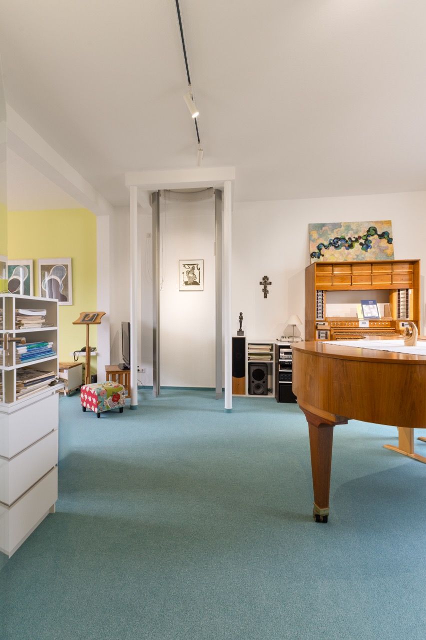 Helles Wohnzimmer mit türkisfarbenem Teppich, Klavier, weißen Wänden und einer Tür, die zu einem anderen Zimmer führt.