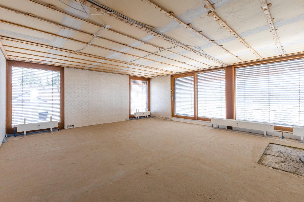 Leerer, unfertiger Raum mit beigem Teppichboden, holzgetäfelter Decke und großen Fenstern, die helles Licht hereinlassen.