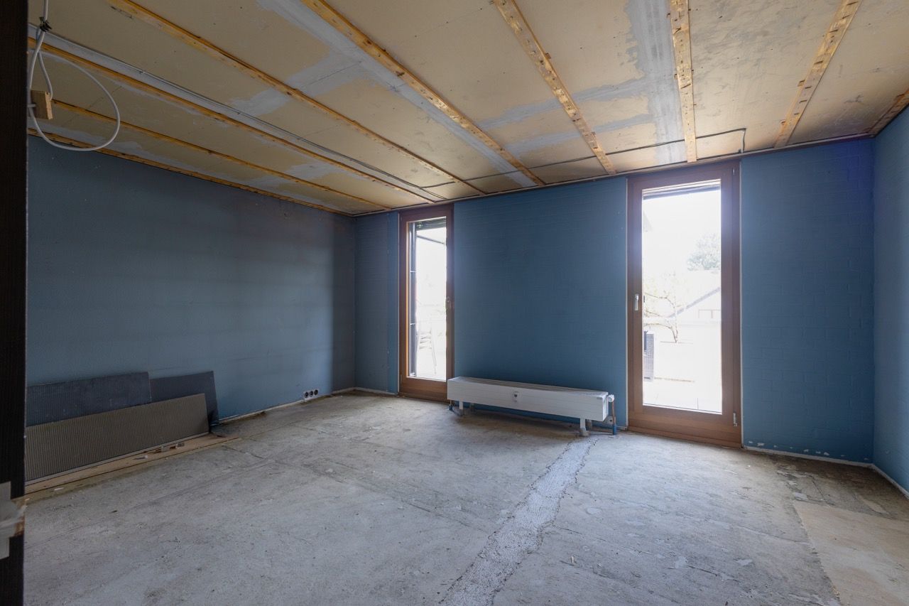 Ein leerer blauer Raum mit unfertigen Wänden, nacktem Boden und zwei hohen Fenstern, wobei sich unter einem der Fenster ein Heizkörper befindet.