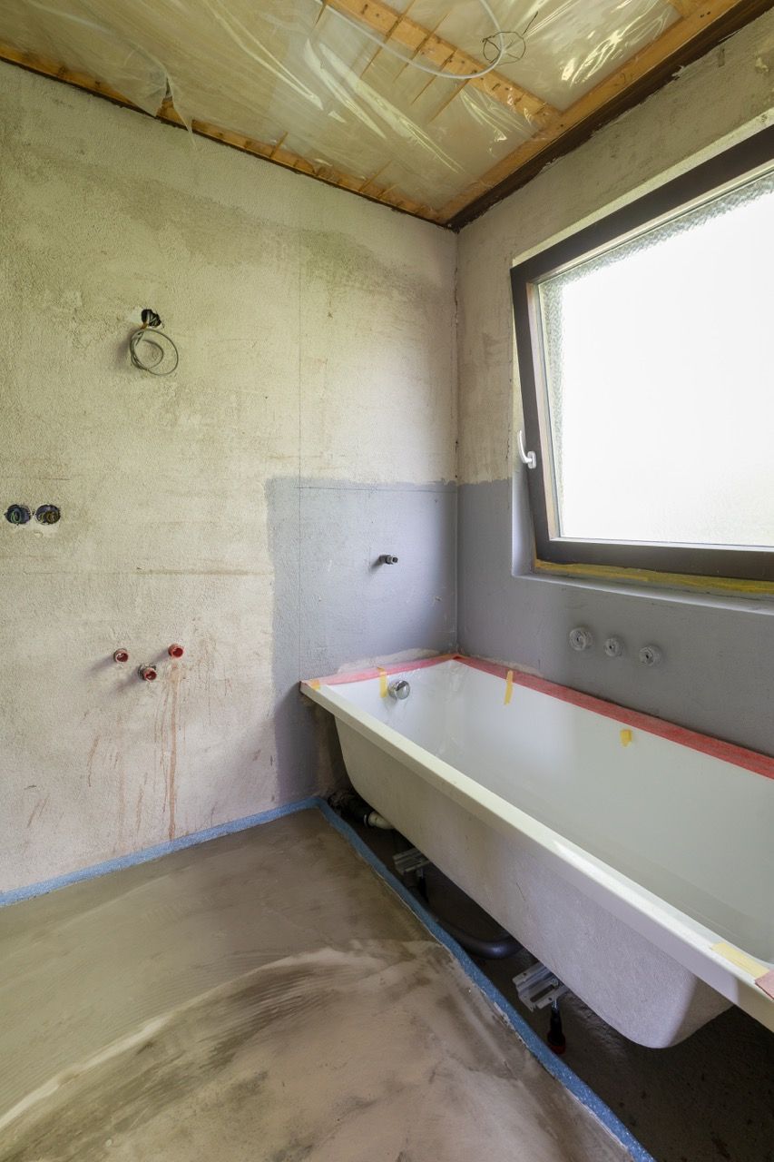 Kleines, noch nicht fertiggestelltes Badezimmer mit weißer Badewanne, Fenster und unverputzten Wänden, das renoviert wird