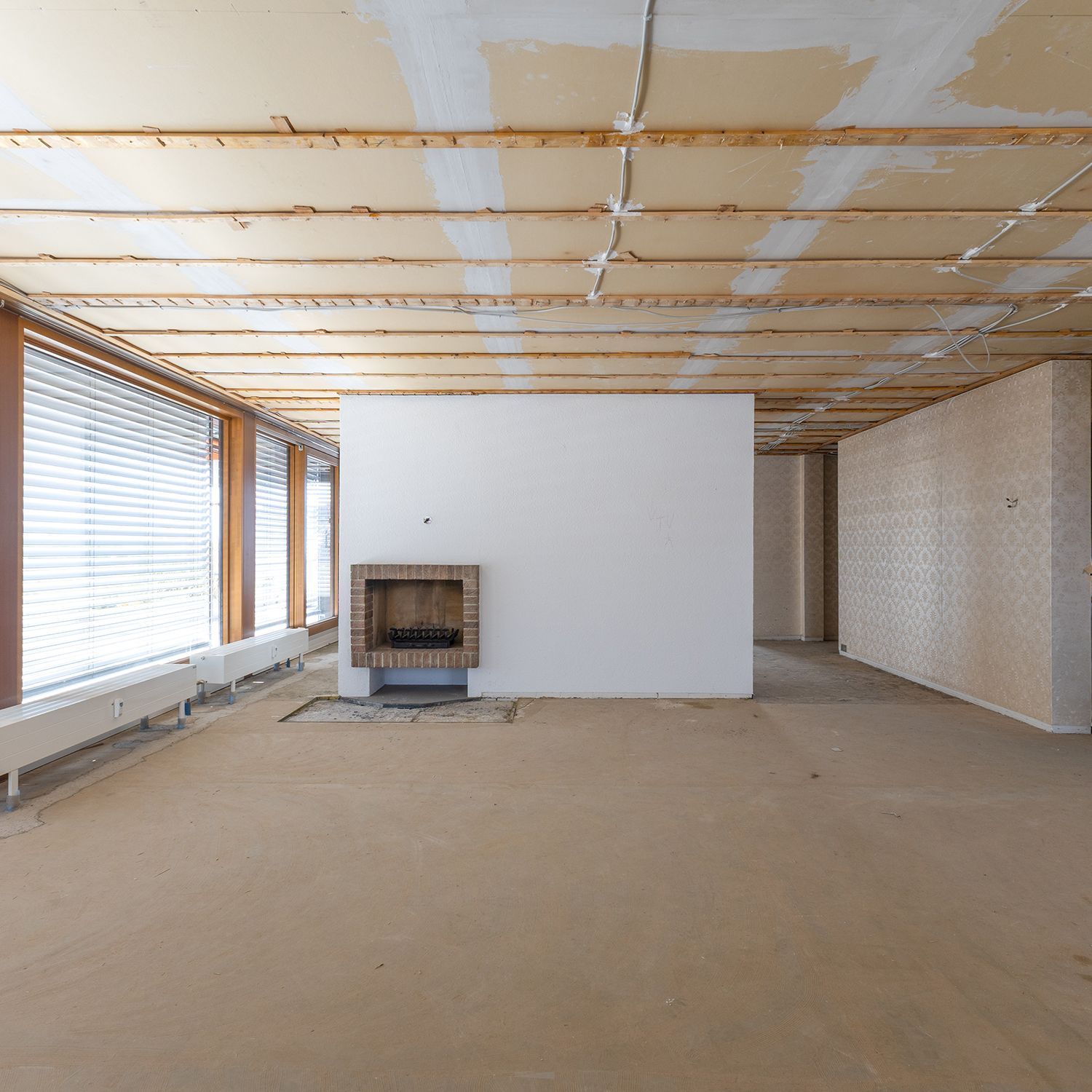Leerer, unfertiger Raum mit freiliegenden Deckenbalken, weißer Trennwand, Kamin und großen Fenstern auf der linken Seite.