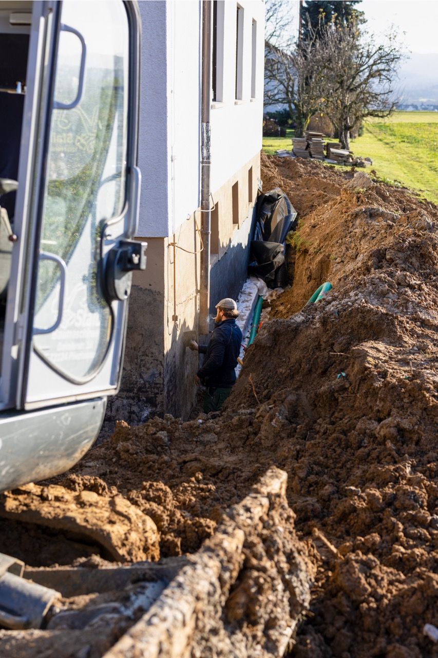 Neben einem weißen Gebäude werden die Fundamente für ein Haus ausgehoben; entlang eines Grabens sind Baggerspuren und aufgeschüttete Erde zu sehen.
