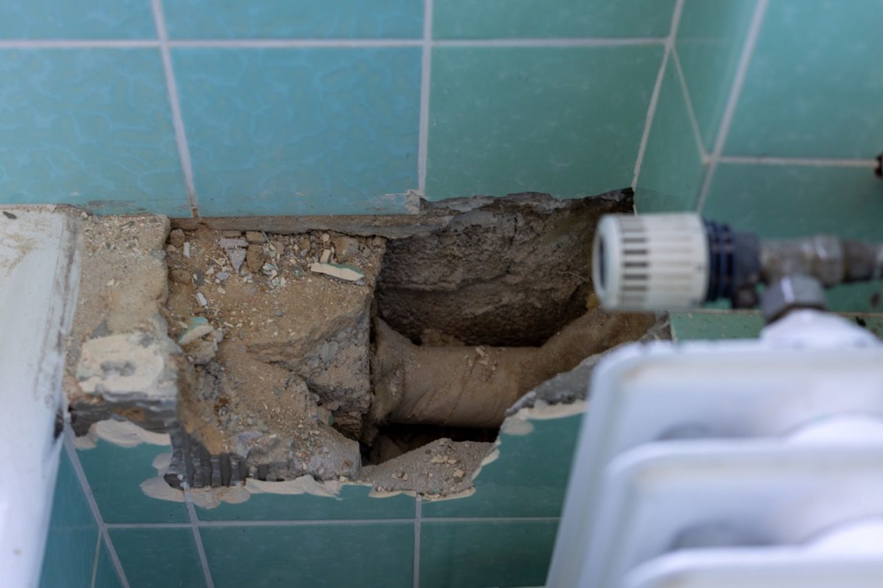 Beschädigte Fliesenwand mit einem großen Loch, das Putz und Rohre neben einem Waschbeckenhahn freilegt