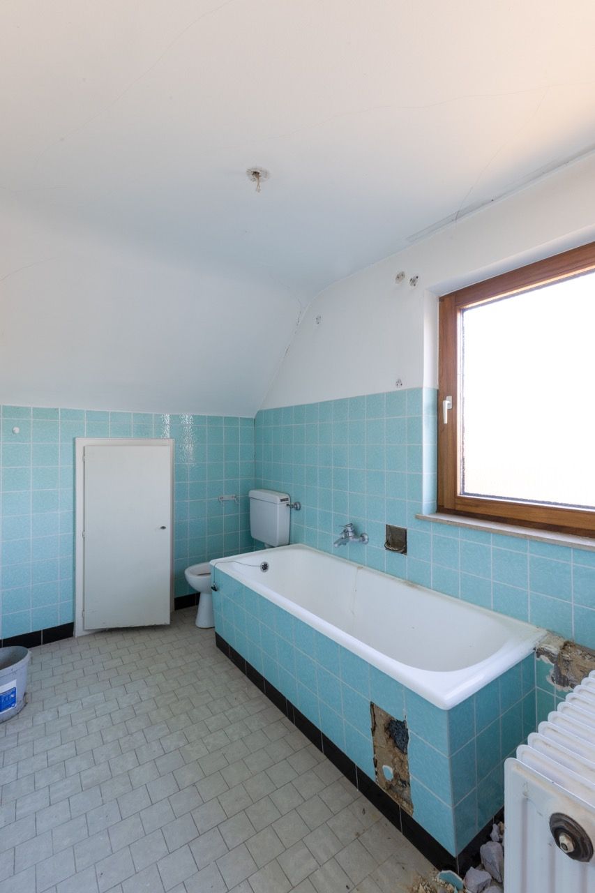 Kleines Badezimmer mit blauen Fliesen, weißer Badewanne, Toilette, Waschbecken und einem Fenster unter einer Dachschräge