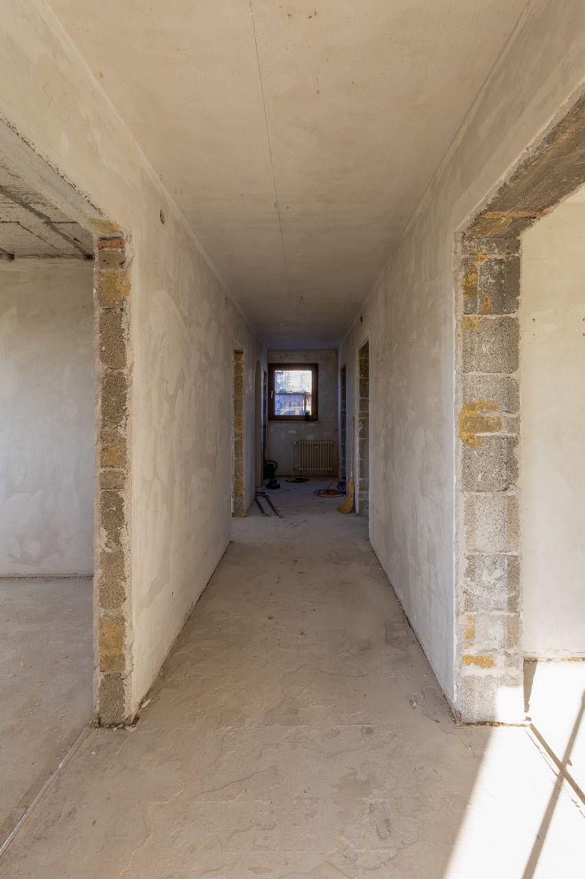 Schmaler, unfertiger Flur in einem im Bau befindlichen Gebäude, mit kahlen Wänden und einem kleinen Fenster am Ende.
