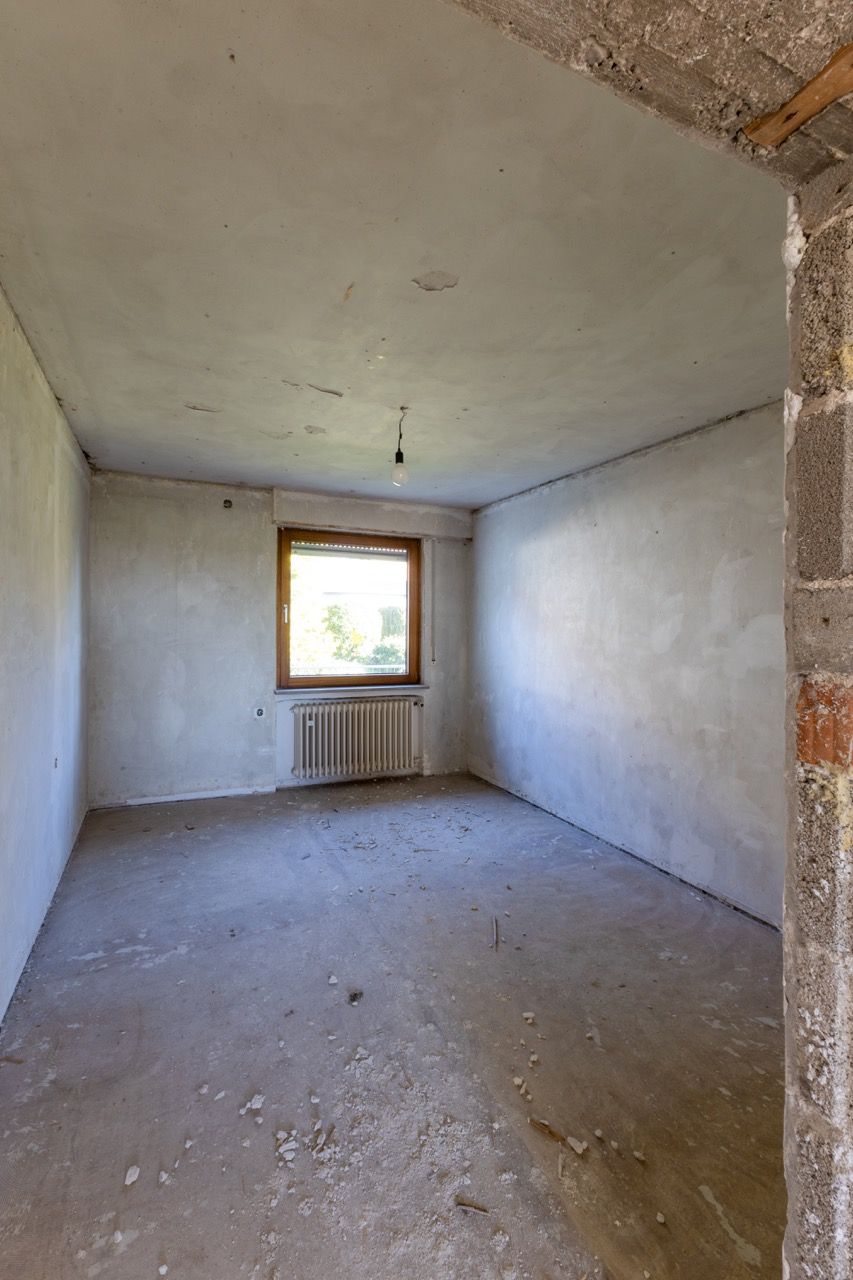 Leerer, unfertiger Raum mit unverputzten Wänden, Betonboden und einem Fenster über einem Heizkörper.