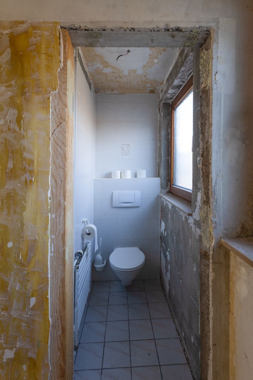 Schmale, unfertige Badezimmer mit Toilette, unverputzten Wänden und einem kleinen Fenster.