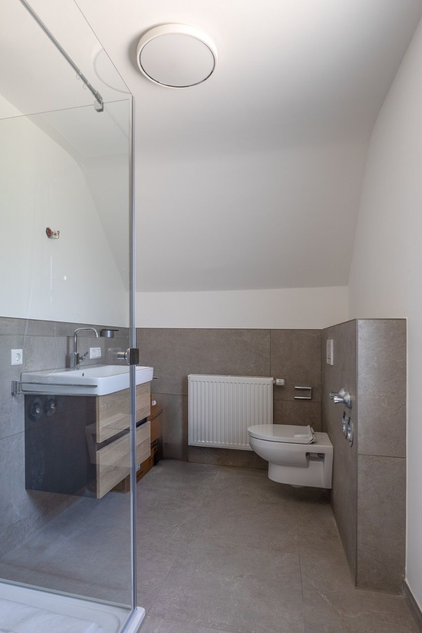 Modernes Badezimmer mit Glasdusche, Waschbecken, Toilette, Heizkörper und grauen Fliesen an Wänden und Boden.