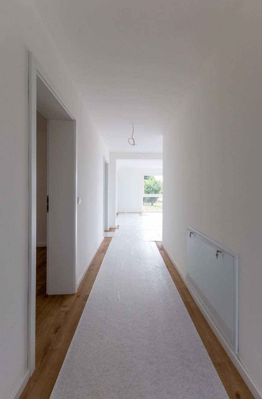 Heller Flur mit weißen Wänden, Holzfußbodenkanten und einem langen Teppichläufer, der zu einem hellen Eingangsbereich führt.