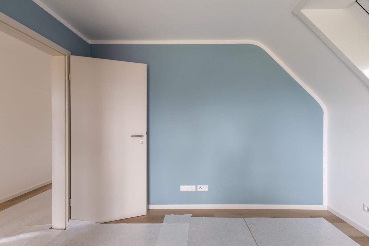 Leerer Raum mit hellblauer Wand, offener weißer Tür und schräger Decke.