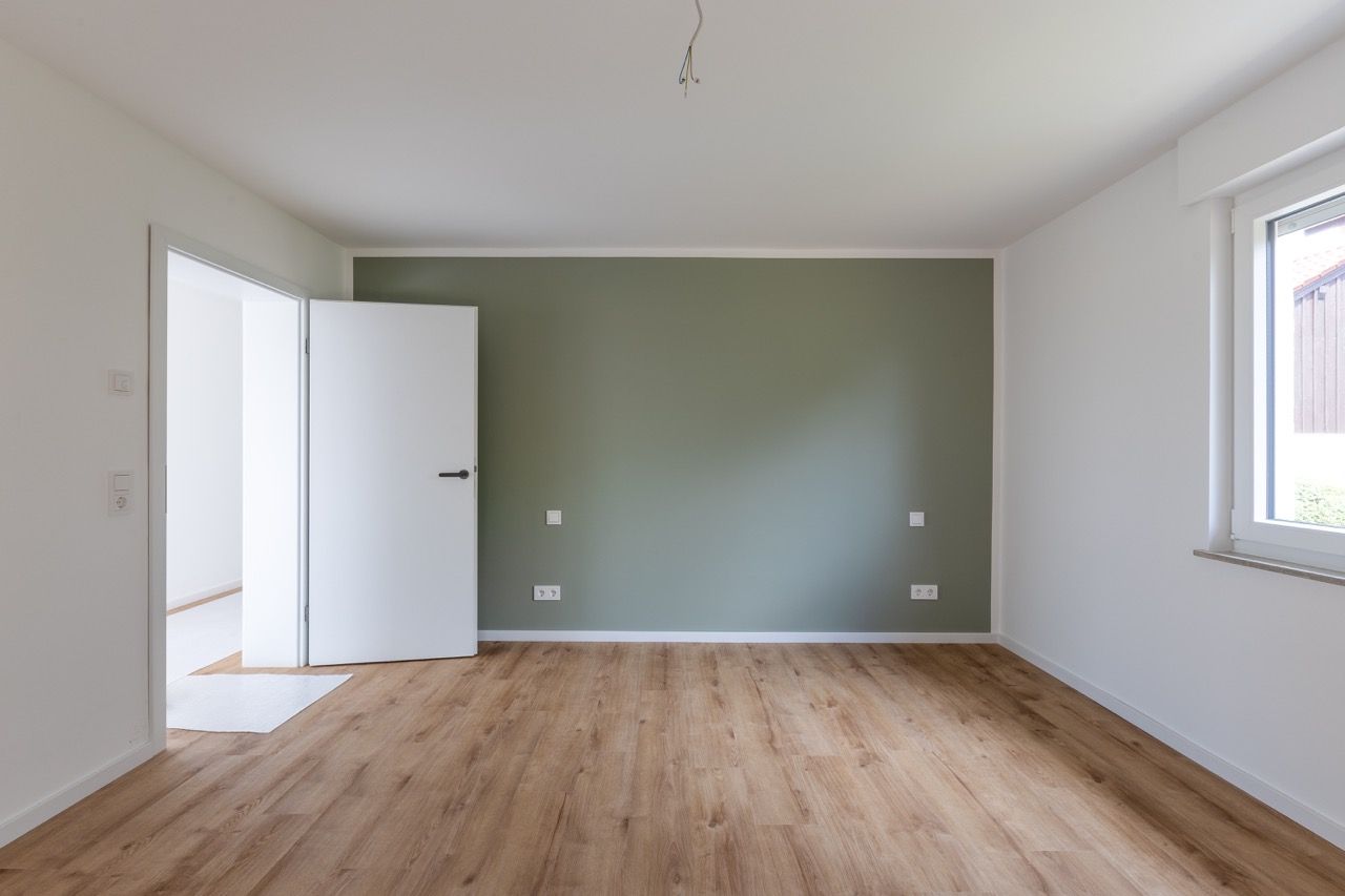 Leerer Raum mit Holzboden, grüner Akzentwand, offener Tür und einem Fenster auf der rechten Seite.