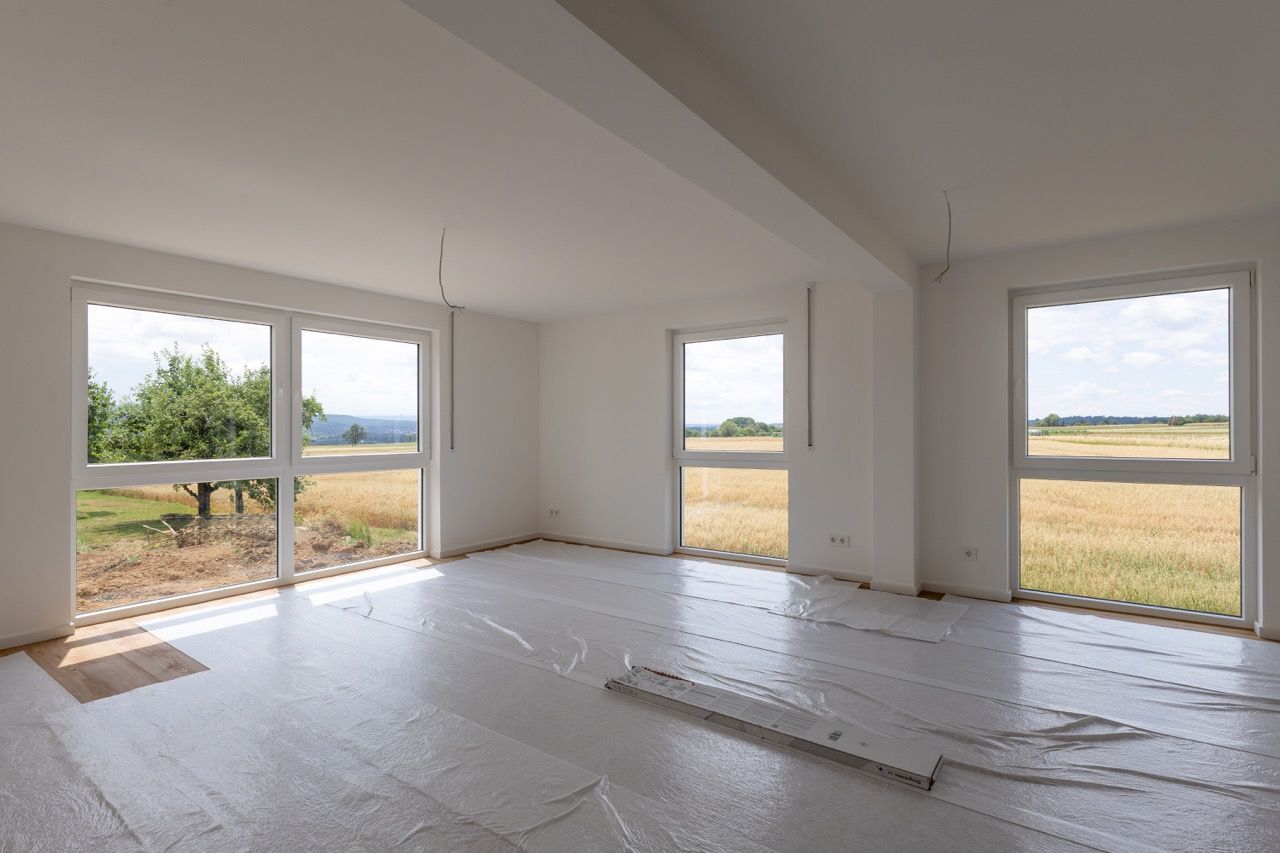 Leerer, unfertiger Raum mit weißen Wänden, freiliegendem Dielenboden und großen Fenstern mit Blick auf ein Feld.