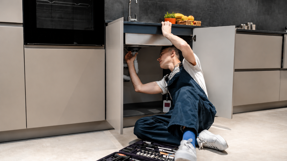 Una persona è inginocchiata in un mobile da cucina, allunga la mano verso l'alto per prendere una torcia e degli attrezzi che si trovano sul pavimento.