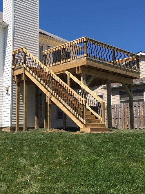 Wooden deck with stairs, black railings, attached to a house with a green lawn and clear blue sky.