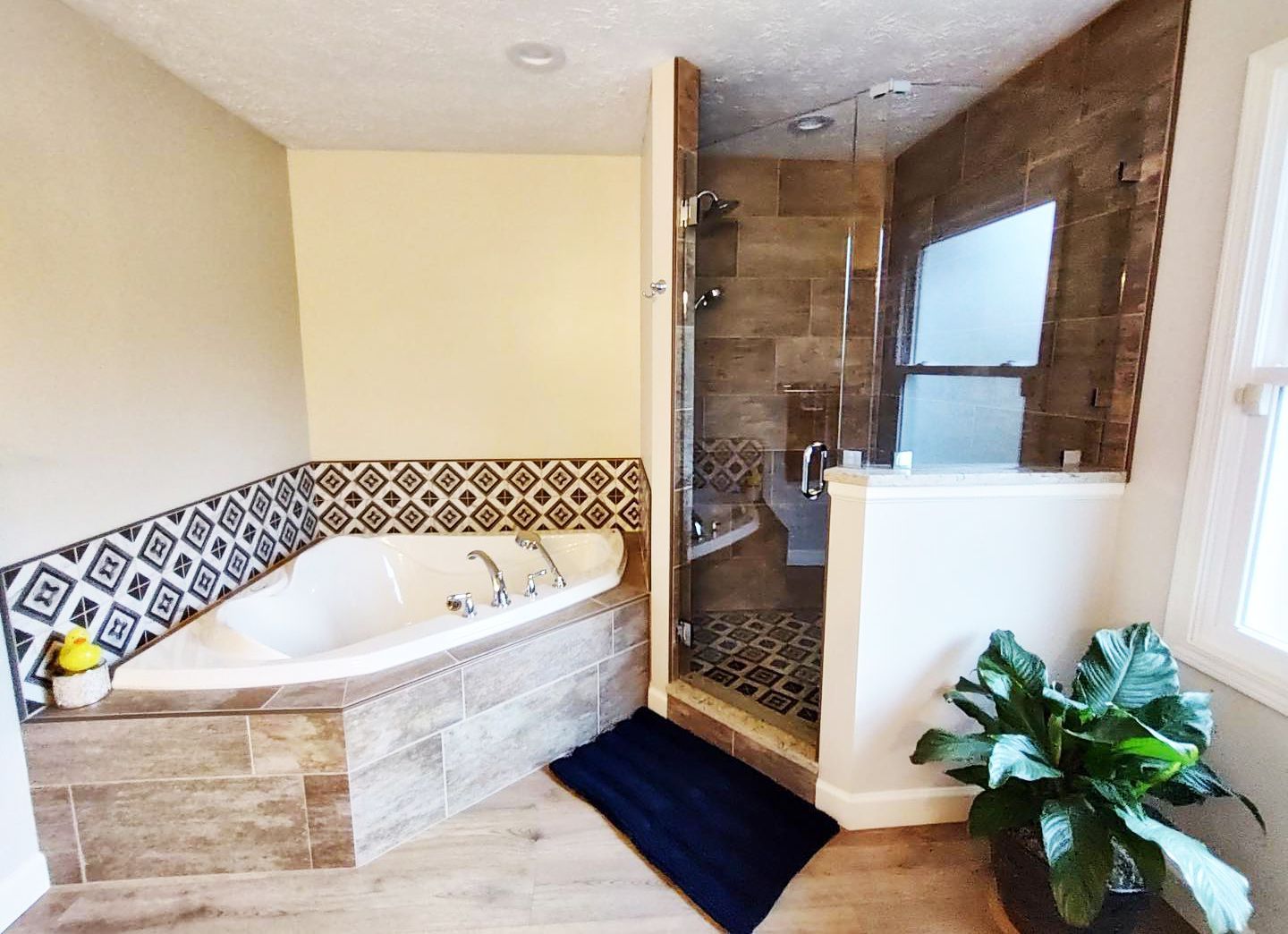 Bathroom with a corner tub, glass shower, and large plant. Beige, brown, and blue tones.