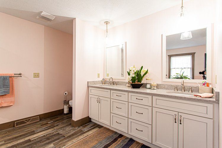 Pink-walled bathroom with light gray double vanity, mirrors, and wooden floor. A window is visible.