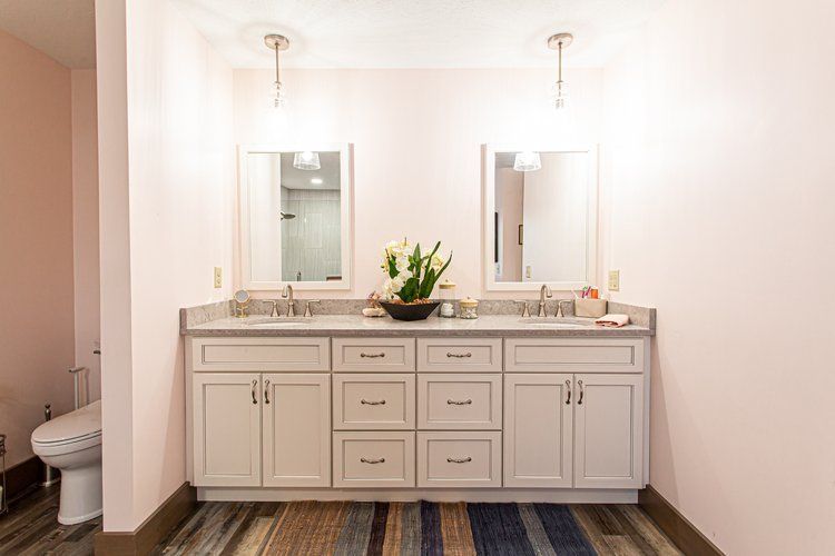 Bathroom with double sinks, two mirrors, and pink walls. Light-colored cabinets, wood-look floor, and pendant lights.