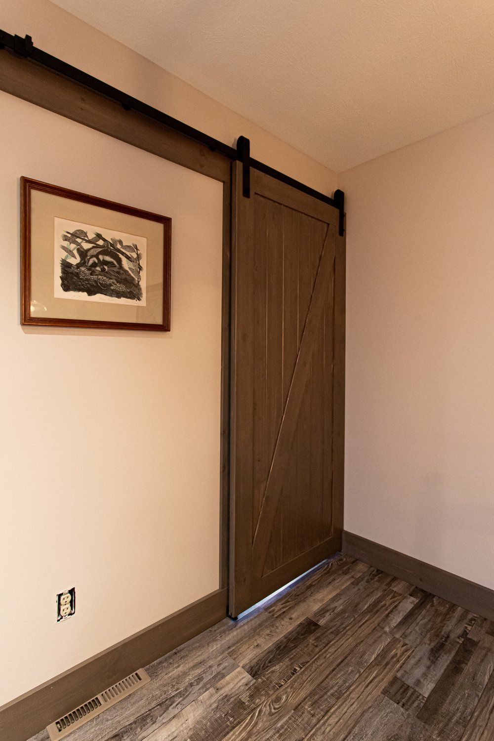Barn door with black hardware, next to a framed print, against beige walls. Dark wood flooring.