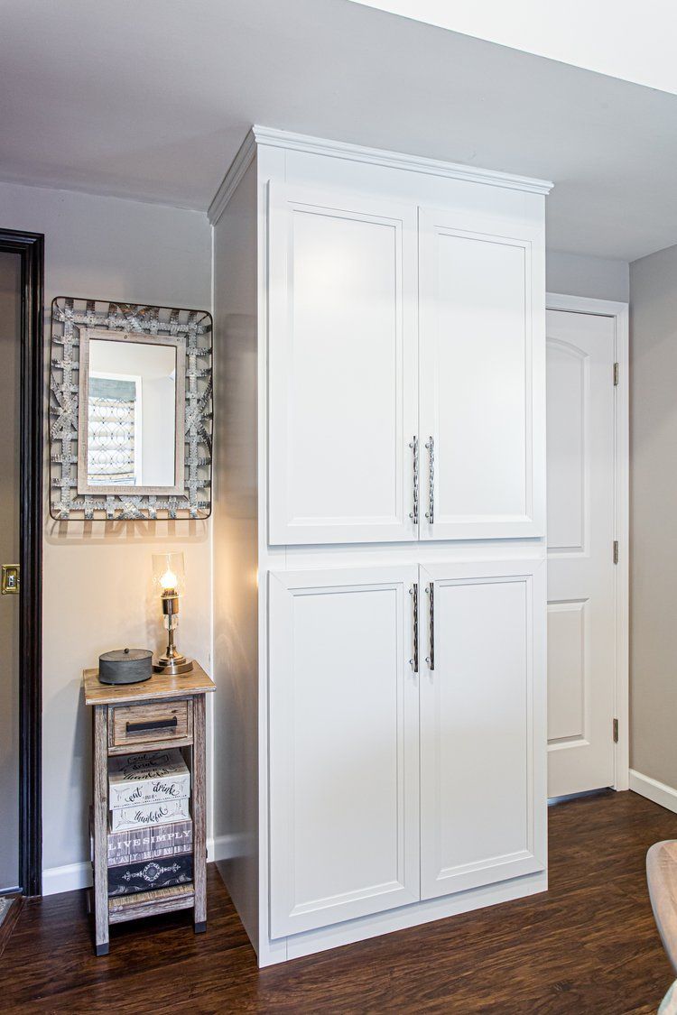 Tall white storage cabinet next to a small wooden side table with a mirror and lamp.