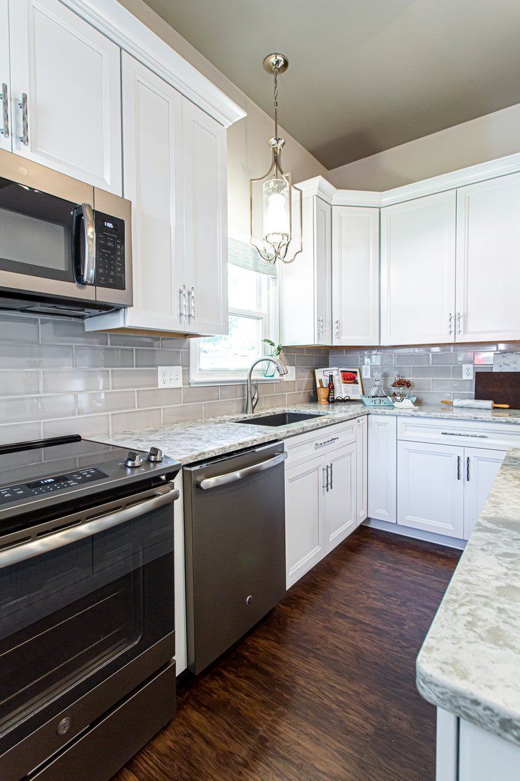 White kitchen with stainless steel appliances, white cabinets, light gray backsplash, and dark wood floors.