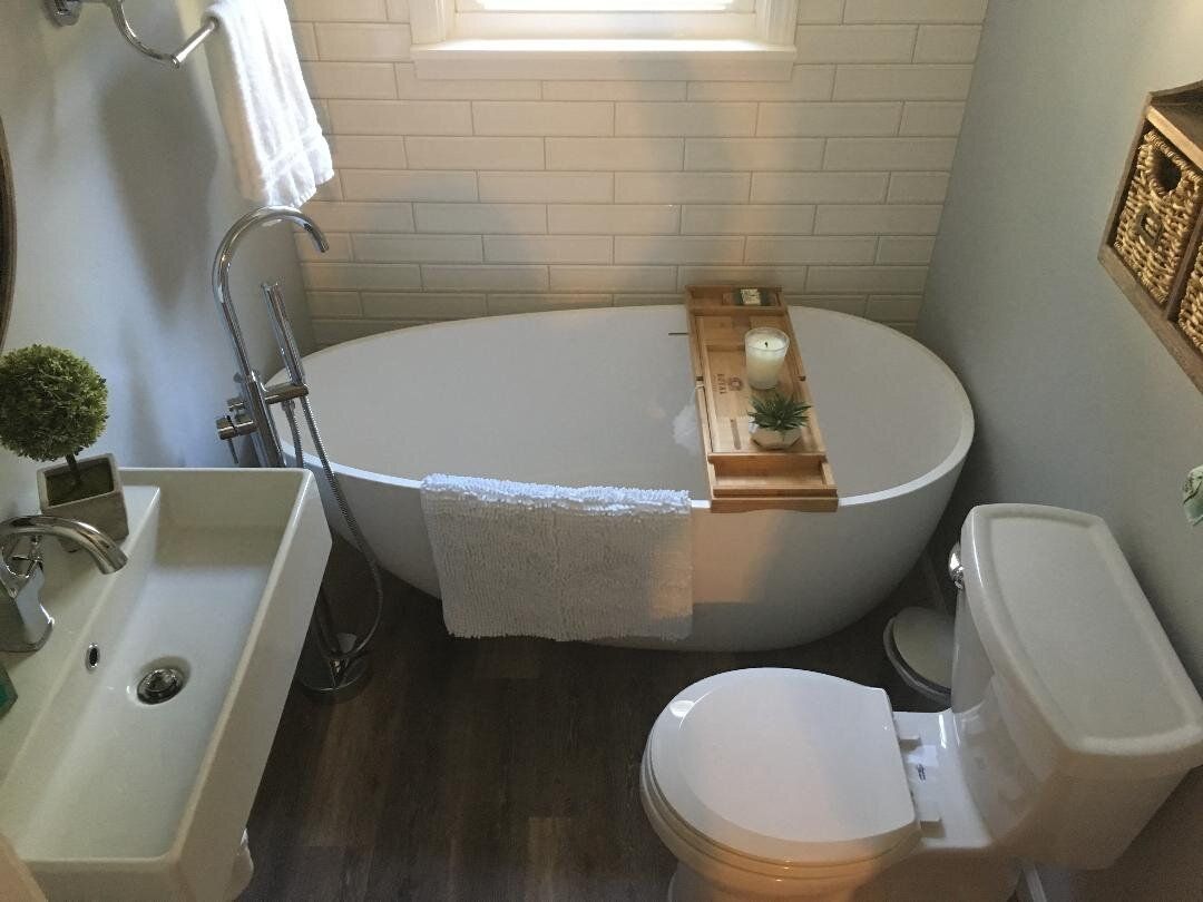 Bathroom with a white oval bathtub, toilet, and sink; wooden floor, tiled wall, and natural light.