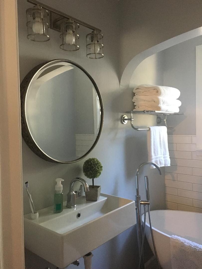 Bathroom with a round mirror, sink, tub, and stacked towels; light gray walls, white fixtures.